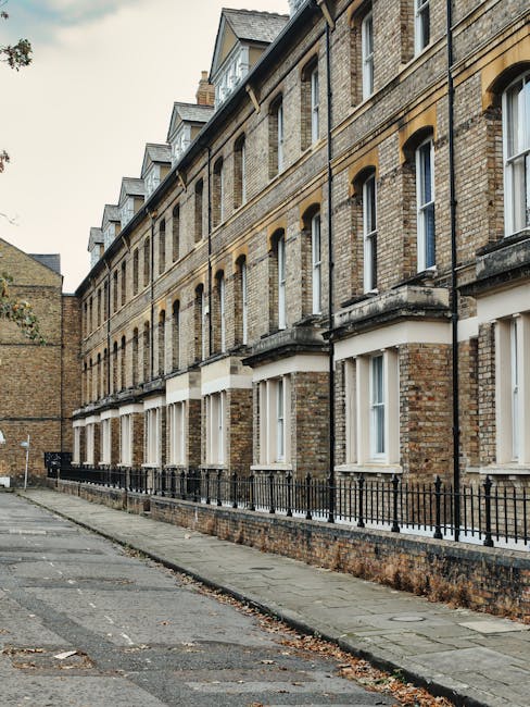 Exterior view of a row of multi-story residential buildings constructed from brick with cream window frames and decorative stone elements, situated along a paved sidewalk. The buildings feature multip